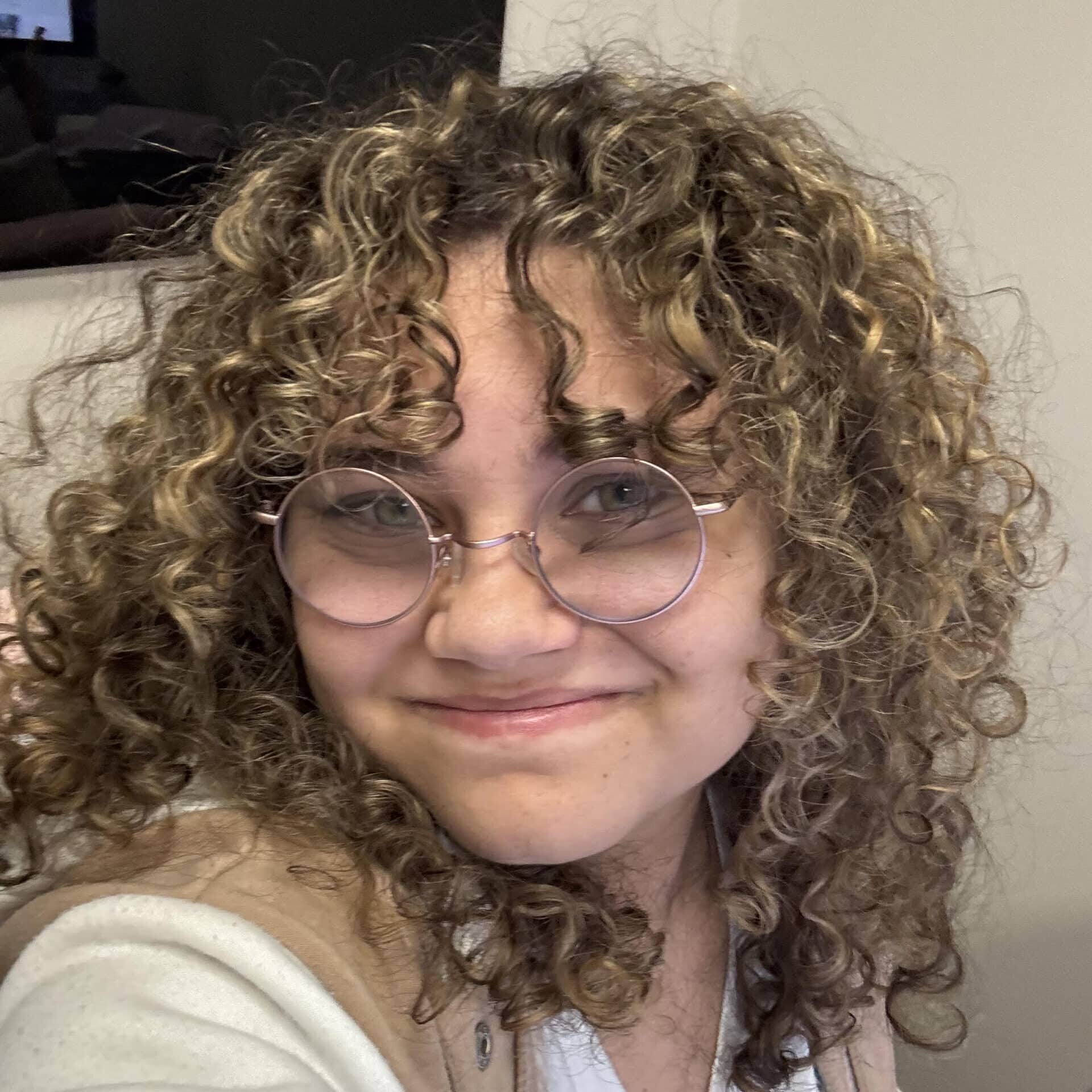 Amidst the glow of ambition's fiery rise, a person with curly hair and round glasses smiles at the camera, embodying the rebirth of creativity through their ambidextrous skills. Clad in a light-colored top and vest, they emerge from behind the screen's digital ashes, where a computer monitor on a desk flickers with hints of their versatile services.